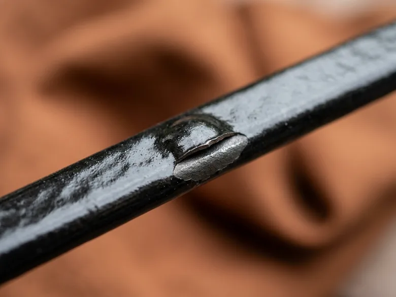 Gros plan macro sur une barre métallique au revêtement noir brillant, dont l’émail est fendu et écaillé, laissant apparaître le métal nu, sur un fond flou de tissu orangé
